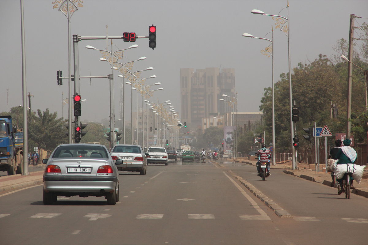 Wide city street with cars, motorbikes and a cyclist under traffic lights and lamp posts, hazy sky