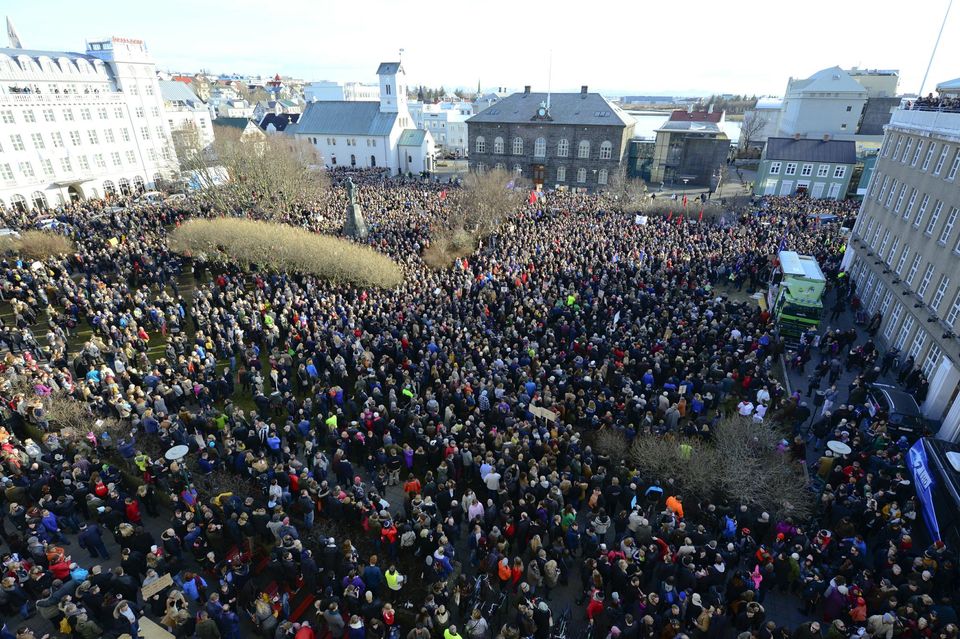 Large crowd of people gathered in a city square surrounded by historic buildings under a bright sky